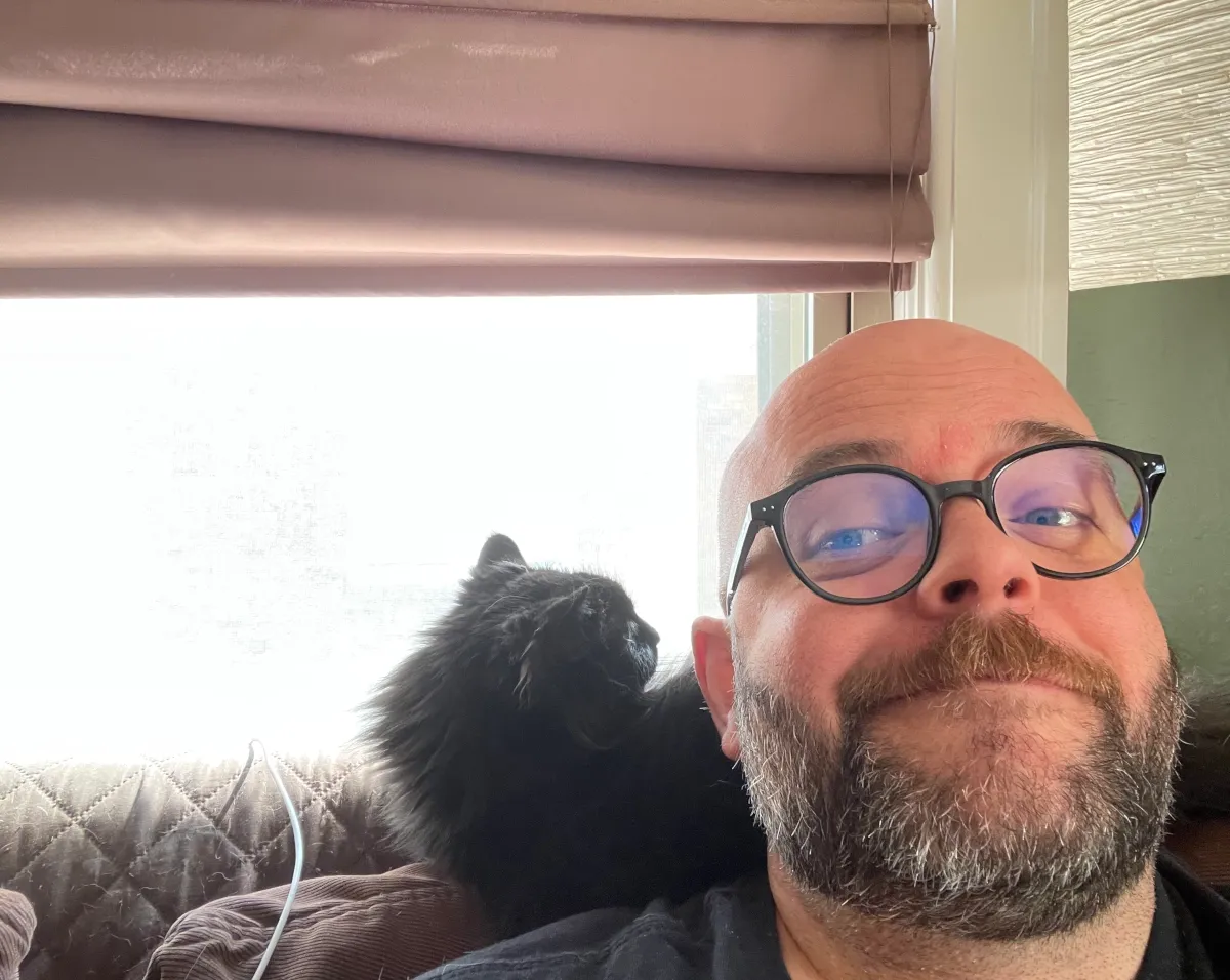 A person with glasses and a beard is smiling at the camera, with a black cat sitting behind them on a couch, looking out a window. Soft light is coming in from the window