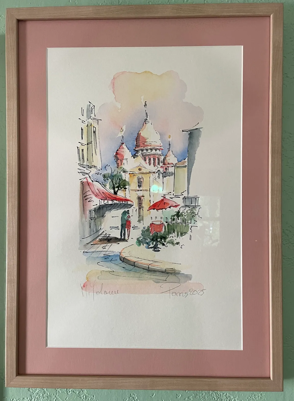 Painting on a light green wall in a light brown frame, pink mat, and featuring Montmartre, France and Sacré-Cœur in the background